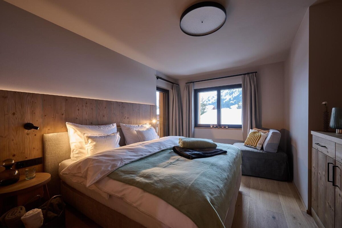 A bedroom with a large bed, a bedside table, a window with curtains, and a wooden headboard.