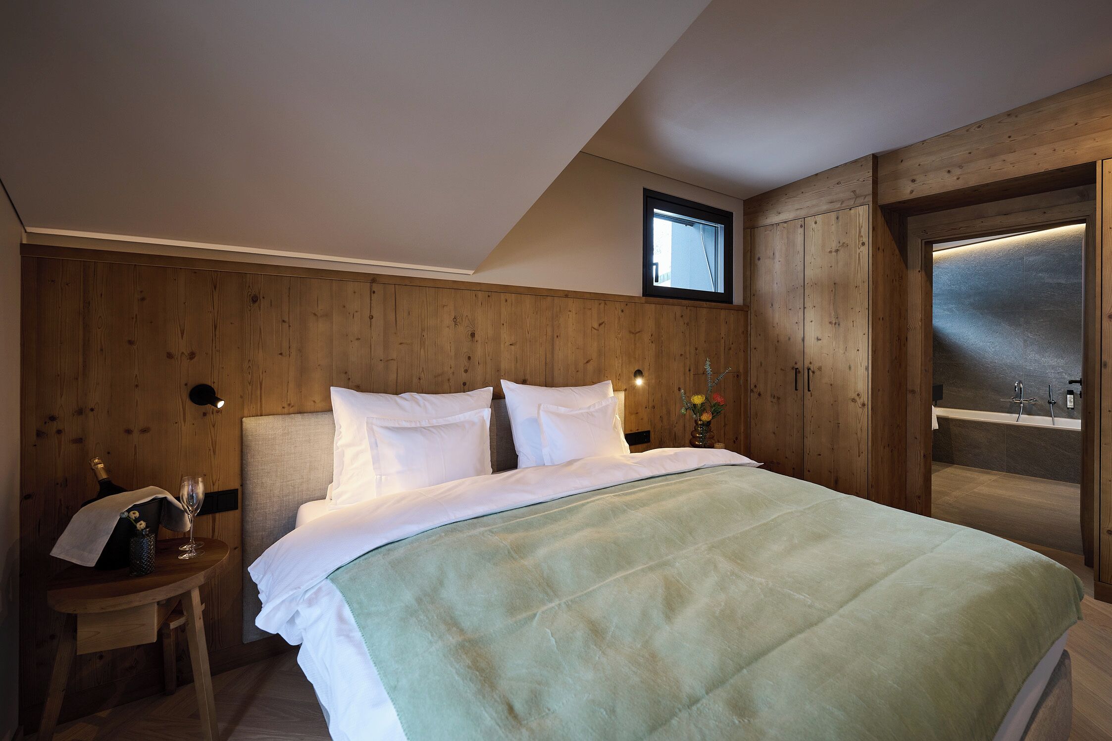 A bedroom with a large bed, wooden panel walls, a small bedside table with a lamp, and a view of a bathroom.