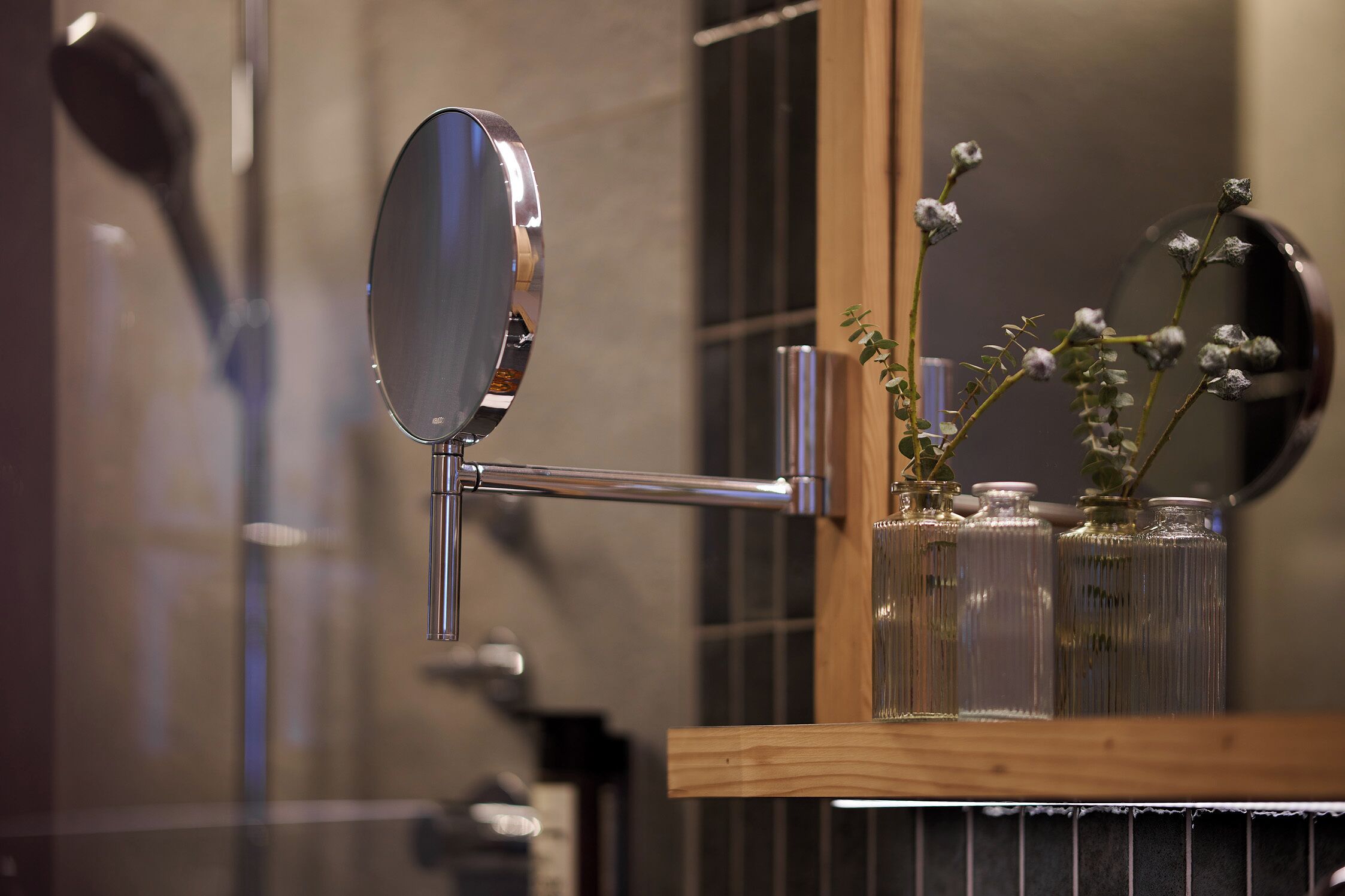 A bathroom with a mirror mounted on a wall, a wooden shelf with glass jars containing dried flowers, and a towel rack.