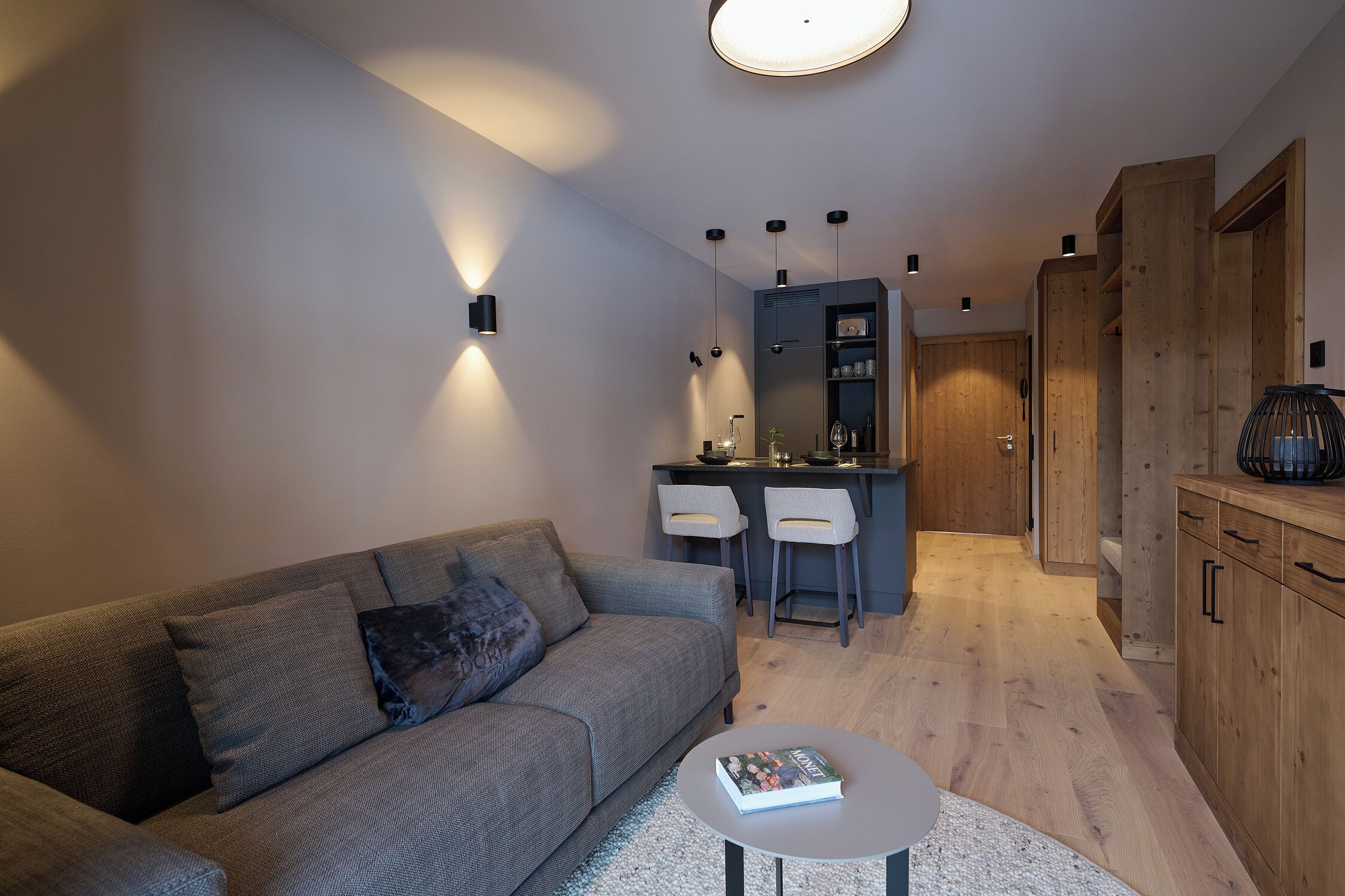 Un salón moderno con un sofá gris, una mesita redonda pequeña y una zona de cocina con taburetes altos y armarios de madera.