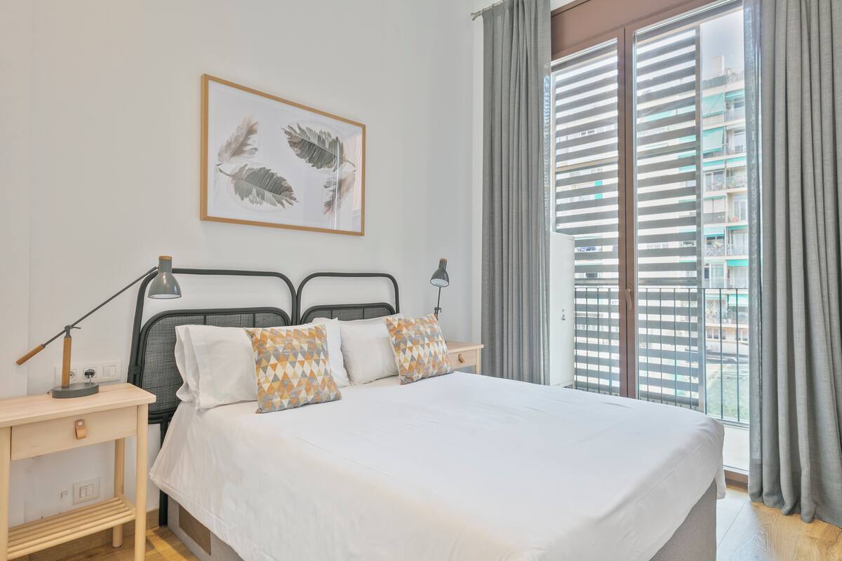 Une chambre à coucher comprenant un lit, deux tables de chevet, une commode, une œuvre d’art encadrée et une fenêtre avec des stores.