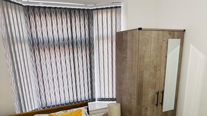 A neatly made bed with a floral bedspread and two yellow pillows, a wooden wardrobe, a bedside table with a lamp, and a window with striped curtains. - Chaucer House Retreat - Leicester (Leicester)