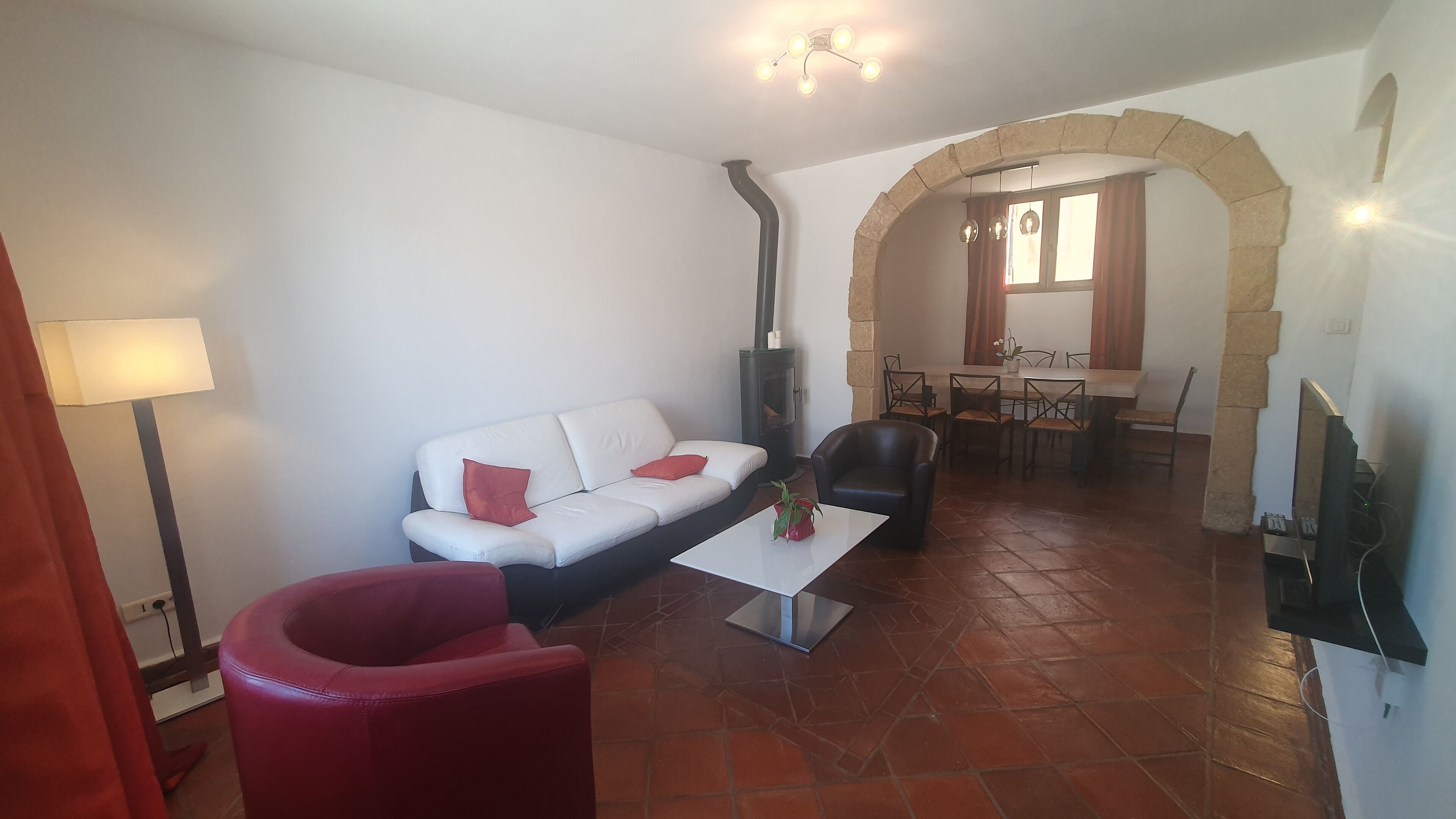 A living room with a white sofa, red armchair, and a coffee table. There is a fireplace, a dining area with a table and chairs, and a stone archway.