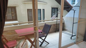 A balcony with a table and chairs, a fan, and a view of a building with barred windows. - Room in Apartment - Jomtien Hill Resort Pratamnak 105 (Pattaya)