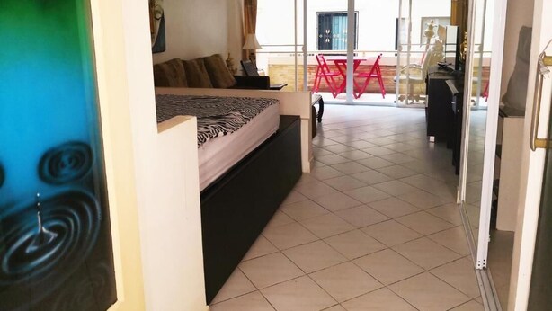 A hotel room with a bed, a sofa, a balcony with red chairs, and a view of the outside. - Room in Apartment - Jomtien Hill Resort Pratamnak 105 (Pattaya)