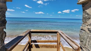 Een houten terras dat uitkomt op een strand met helderblauw water en een stralend blauwe lucht.