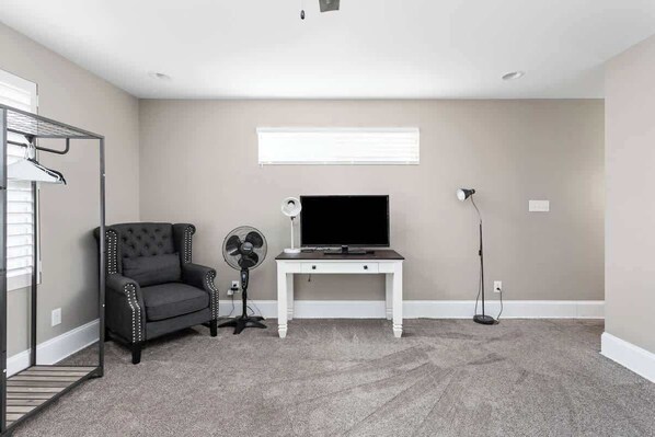 A room with a grey carpet, a white desk with a flat-screen TV, a black upholstered chair, a standing fan, and a floor lamp. - Urban Retreat | Walkable | Easy Parking | Spacious (Indianapolis)