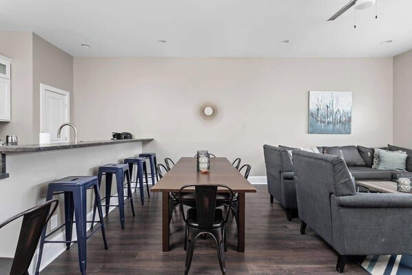 Une cuisine moderne avec un îlot central, un coin repas avec une longue table et des chaises, et un canapé gris dans l’espace salon.