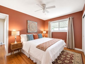 Quarto com cama, duas mesinhas de cabeceira, ventoinha no teto, janela com cortinas e tapete estampado.