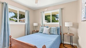 Quarto com cama de madeira, duas mesas de cabeceira com luminárias, ventilador de teto e janelas com cortinas.