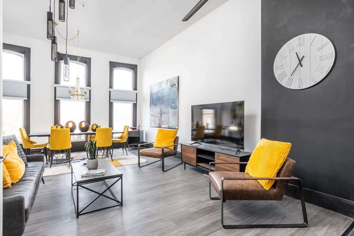 Un salón moderno con un televisor de pantalla plana grande, un reloj redondo en la pared y una zona de comedor con sillas amarillas y una mesa.