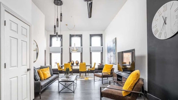 Un salon moderne avec un canapé, des fauteuils, une table basse et un téléviseur. De grandes fenêtres et une horloge ornent le mur.