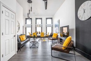 Una sala de estar moderna con un sofá, sillas, una mesa de centro y un televisor. Hay ventanas amplias y un reloj en la pared.