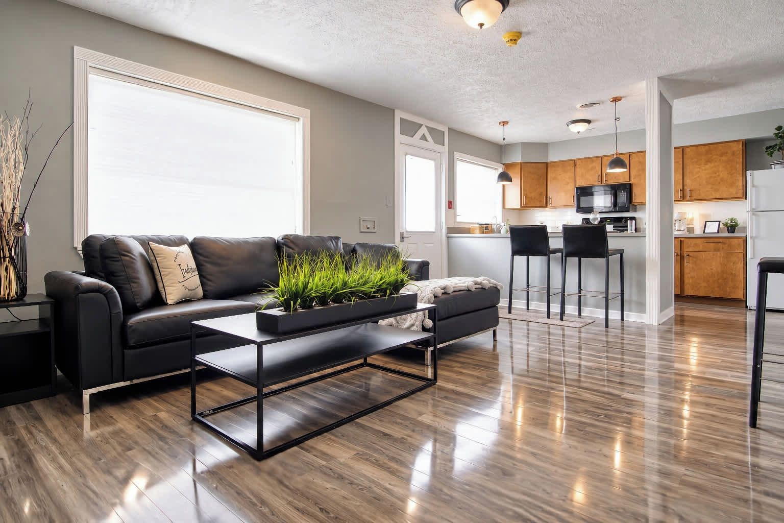 A modern living room with a black leather sofa, a coffee table, and a kitchen in the background.