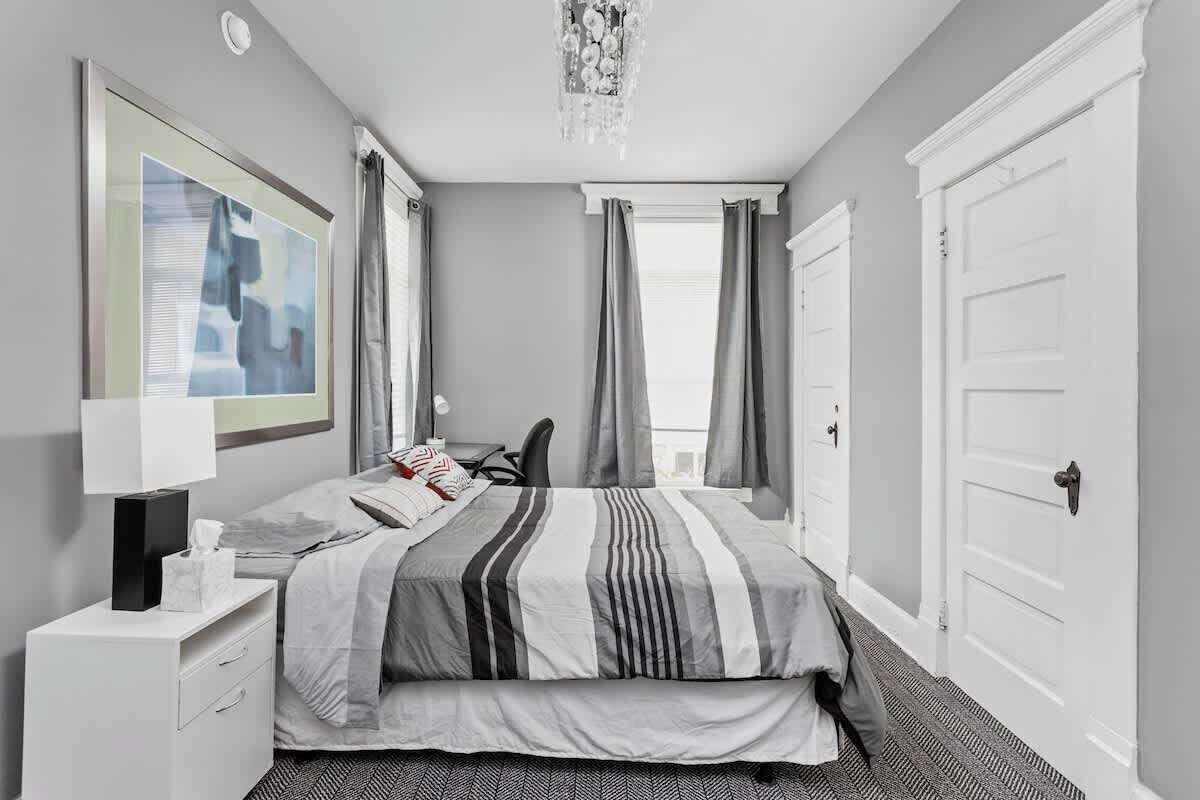 Quarto de hotel moderno com uma cama grande, uma escrivaninha e uma cadeira. Há uma janela com cortinas, um candelabro e um quadro na parede.