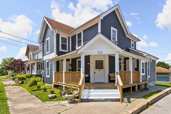 A two-story house with a front porch, a number '215' on the front door, and a small garden with a bench.