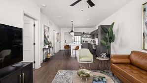 Un salon moderne avec un téléviseur, un canapé en cuir marron, une table basse agrémentée d’objets décoratifs, et un coin repas avec des chaises, le tout avec une cuisine en arrière-plan.