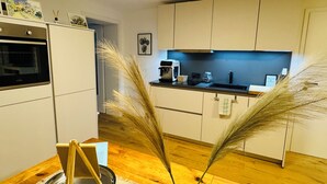 A modern kitchen with white cabinets, a built-in oven, and a coffee maker. There are decorative pampas grass plants in the foreground.