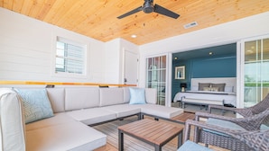 A modern outdoor living area with a wooden ceiling, a white sofa with patterned cushions, a wicker chair, and a wooden table.