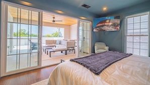 A bedroom with a large bed, a sliding glass door leading to a balcony with outdoor seating, and a ceiling fan.