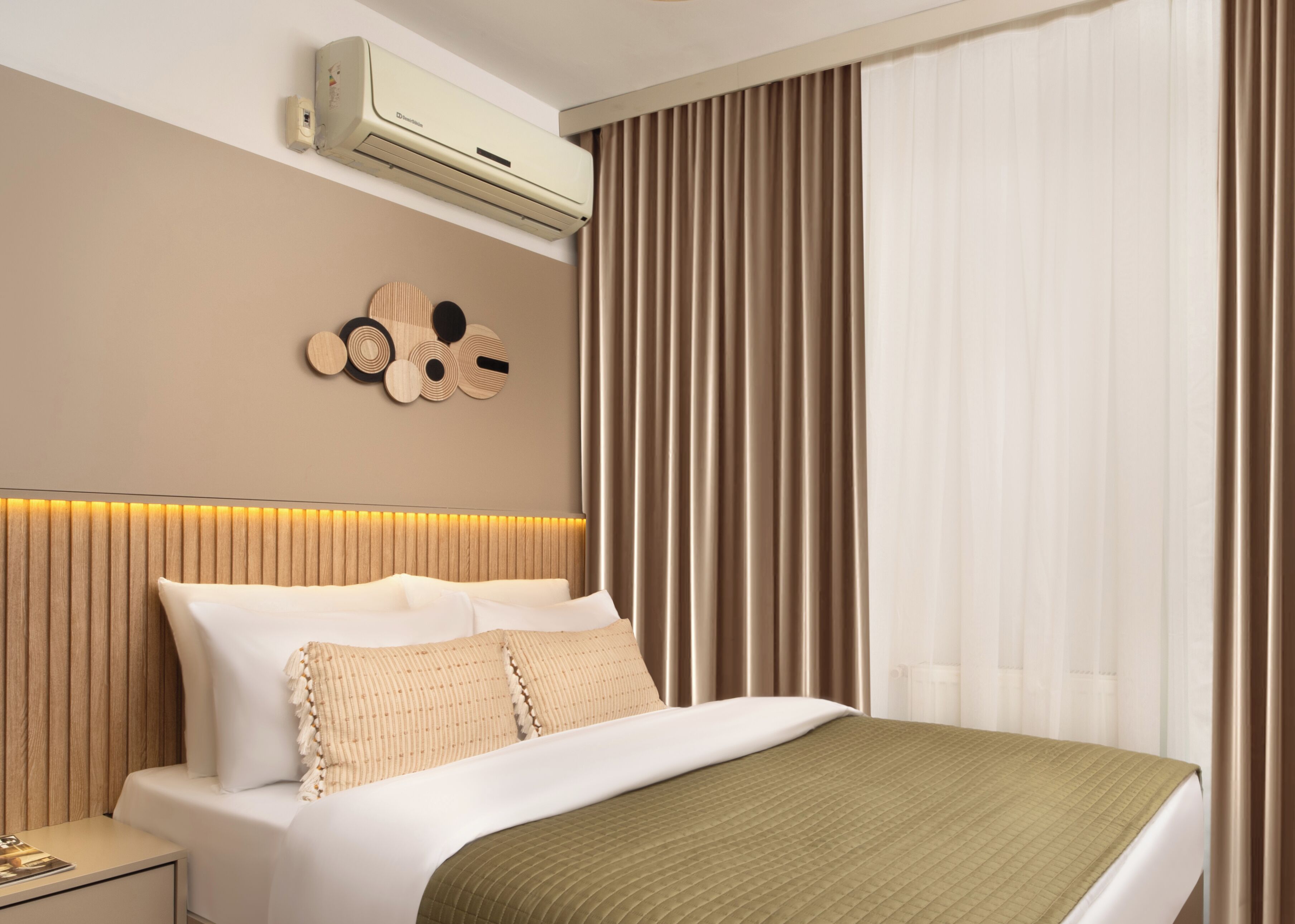 A modern hotel room with a large bed, a wall-mounted air conditioner, a decorative wall piece, and a window with curtains.