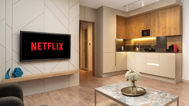 A modern living room with a flat-screen TV displaying "NETFLIX", a kitchen with wooden cabinets, and a dining area with a marble table. - Marmada Suites (Istanbul)