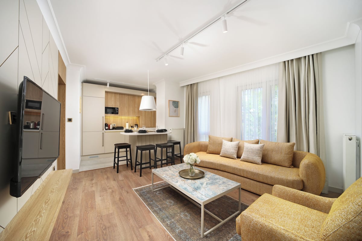 A modern living room with a sofa, coffee table, and TV. There is a kitchen area with a dining set and bar stools.