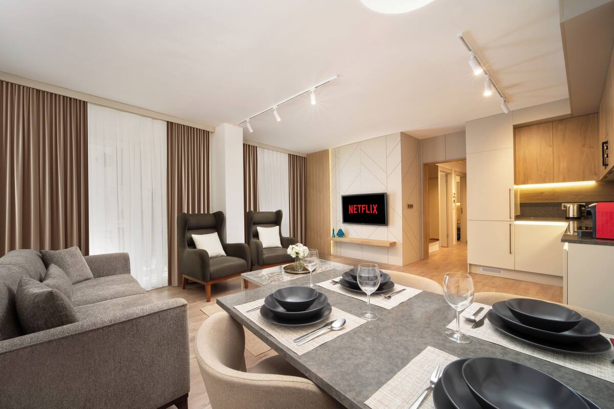 A modern living room with a sofa, armchairs, a dining table set with plates and glasses, and a TV displaying 'NETFLIX'.