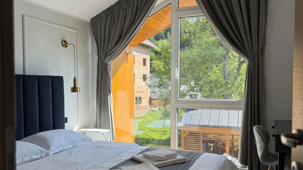 A modern bedroom with a large bed, a desk, and a view of greenery outside.