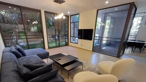 A modern living room with a sofa, a coffee table, and two chairs. There is a TV on the wall and large windows providing a view of the outdoors.