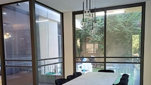 A modern dining area with a large table and chairs, large windows with a view of greenery, and a hanging light fixture.