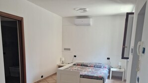 A bedroom with a bed, a sofa, a bedside table, a wall-mounted air conditioner, and a door leading to another room.