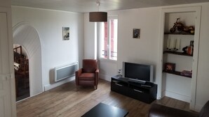 A living room with a brown leather chair, a black entertainment unit with a TV, wooden flooring, and a window with a view of a red building.