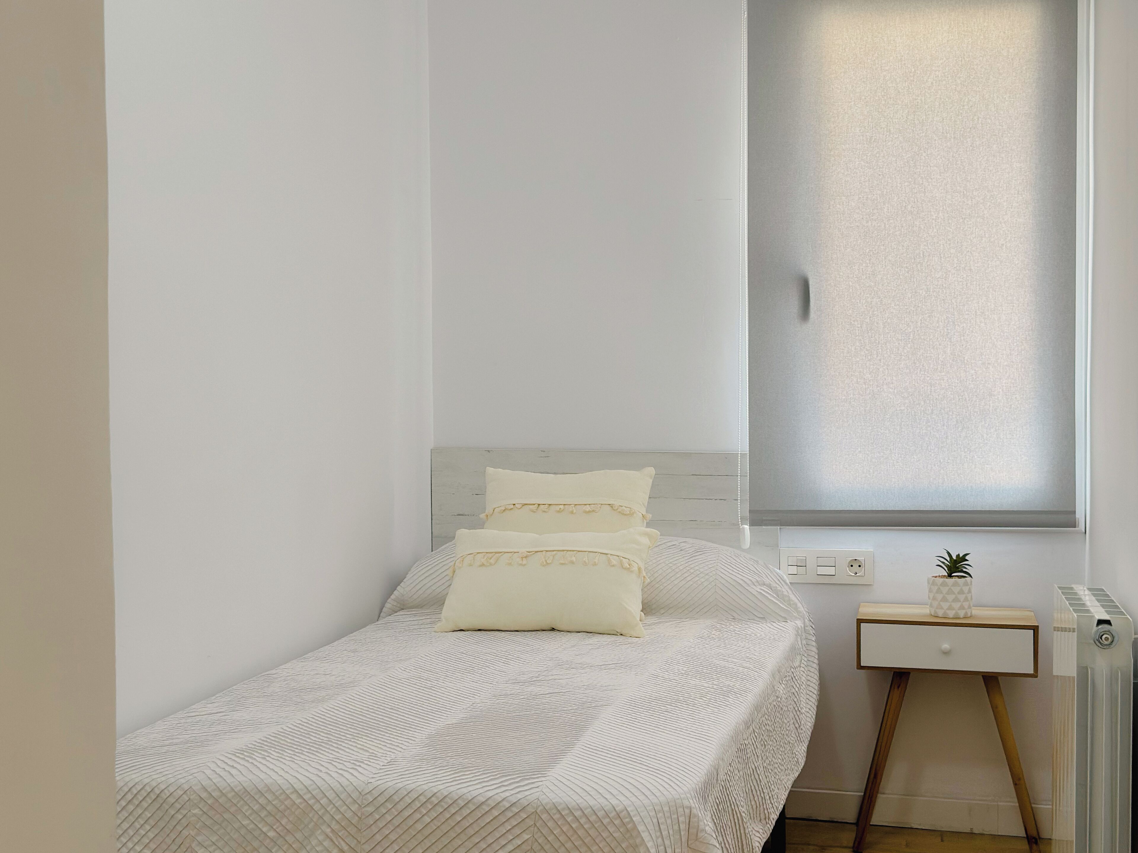 A small, neatly arranged bedroom with a bed, a bedside table, a radiator, and a window with blinds.