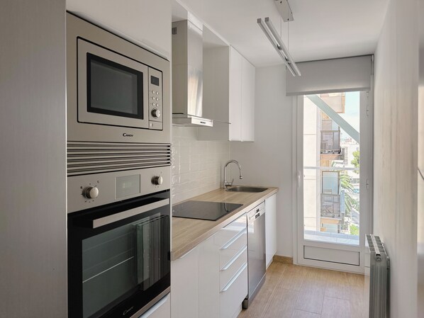 A modern kitchen with a built-in microwave, oven, and dishwasher. It features white cabinetry, a wooden countertop, and a window providing a view of the outside. - APARTAMENTO RENOVADO A 10 M DE LA PLAYA CON VISTAS AL MAR (Salou)