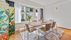 A modern dining area with a wooden table and chairs, a large window with curtains, and a view of buildings outside. - Central & luminous apartment in Antwerp, foreseen with all comforts (Antwerpen)