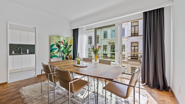A modern dining area with a wooden table and chairs, a white rug, and a view of a building through large windows. - Central & luminous apartment in Antwerp, foreseen with all comforts (Antwerpen)