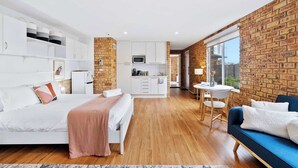 A modern bedroom with a large bed, a sitting area with a blue sofa, a kitchen area, and a brick wall. - 'Hillside Studio' self-contained with City Views. (Howrah)