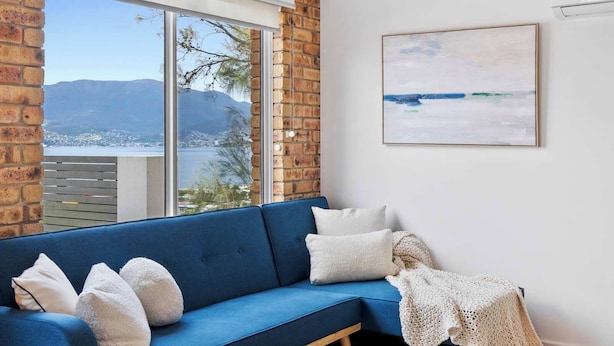 A blue sofa with white cushions, a knitted throw, and a view of a coastal landscape through a large window. - 'Hillside Studio' self-contained with City Views. (Howrah)