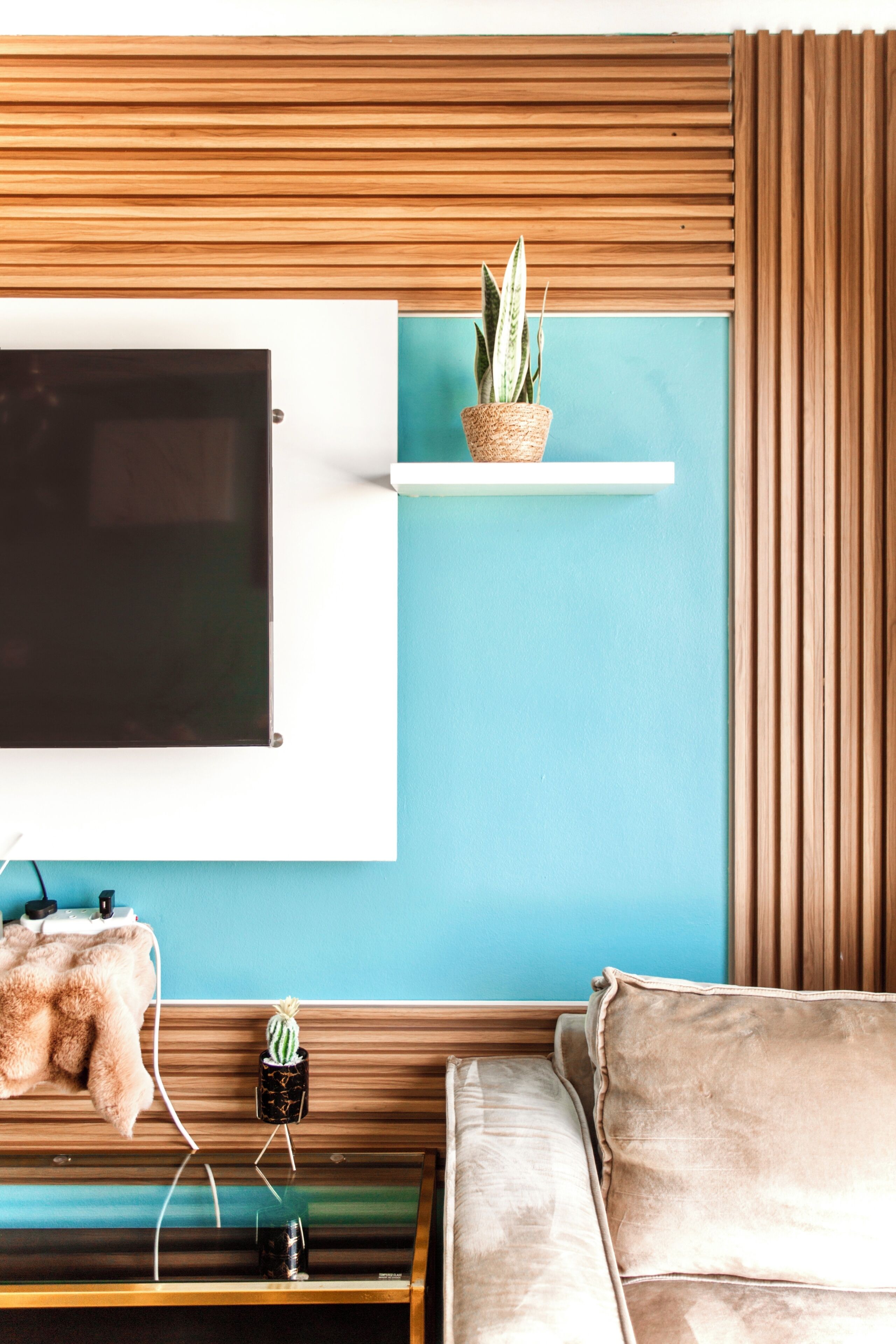 A room with a TV mounted on a wall, a wooden slat design, a teal accent wall, a sofa, and two potted plants.