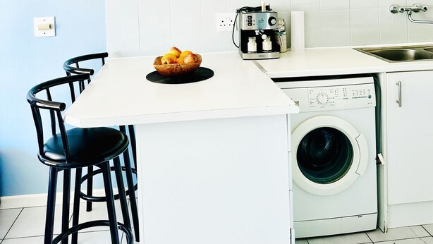 A compact kitchen with white cabinets, a coffee machine, a washing machine, and a small dining area with stools. - Caline VIP Beach Close (Cape Town)