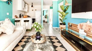 A modern living room with a sofa, coffee table, TV, and kitchen area. - Caline VIP Beach Close (Cape Town)