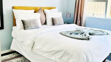 Una cama bien tendida con ropa blanca y un plaid gris, cabecera con un detalle amarillo, un cuadro en la pared y una ventana con cortinas.