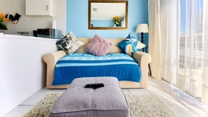 A modern living room with a beige sofa, a blue and white patterned throw, a fluffy pink pillow, and a grey ottoman with a remote control. - Caline VIP Beach Close (Cape Town)
