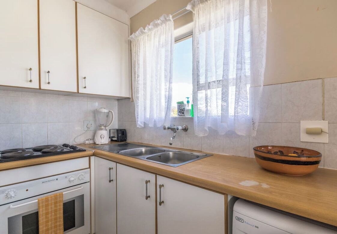 Una cocina con armarios blancos, estufa, lavamanos y una ventana con cortinas traslúcidas.