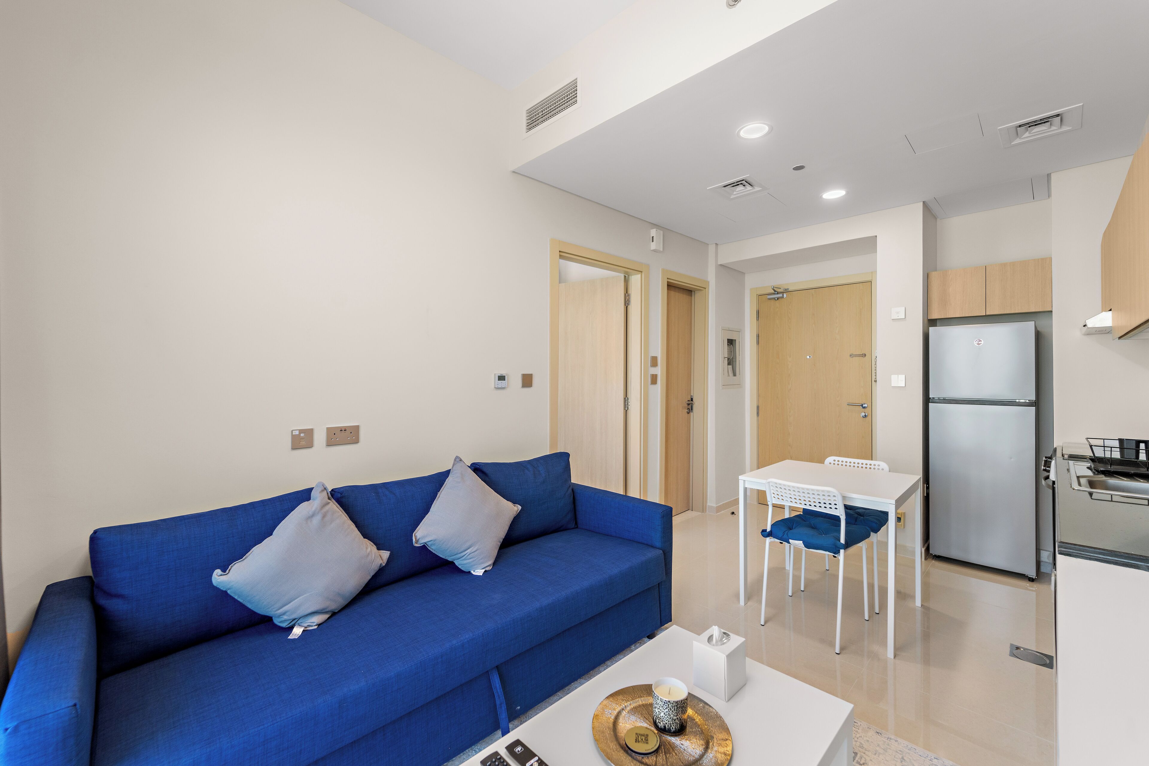 Uma sala de estar moderna com um sofá azul, uma pequena mesa de jantar e uma área de cozinha com geladeira.