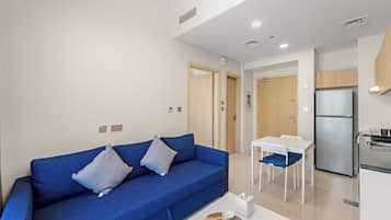 Uma sala de estar moderna com um sofá azul, uma pequena mesa de jantar e uma área de cozinha com geladeira.