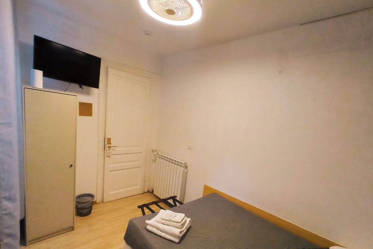 A small, simple room with a bed, a desk, a chair, a radiator, and a television mounted on the wall.