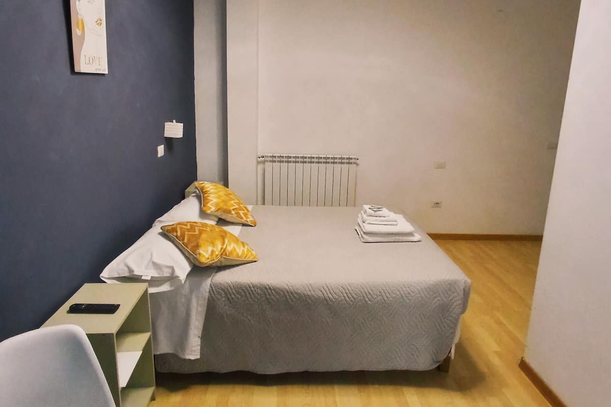 A bedroom with a bed, two pillows, a bedside table, a chair, and a ceiling light fixture.