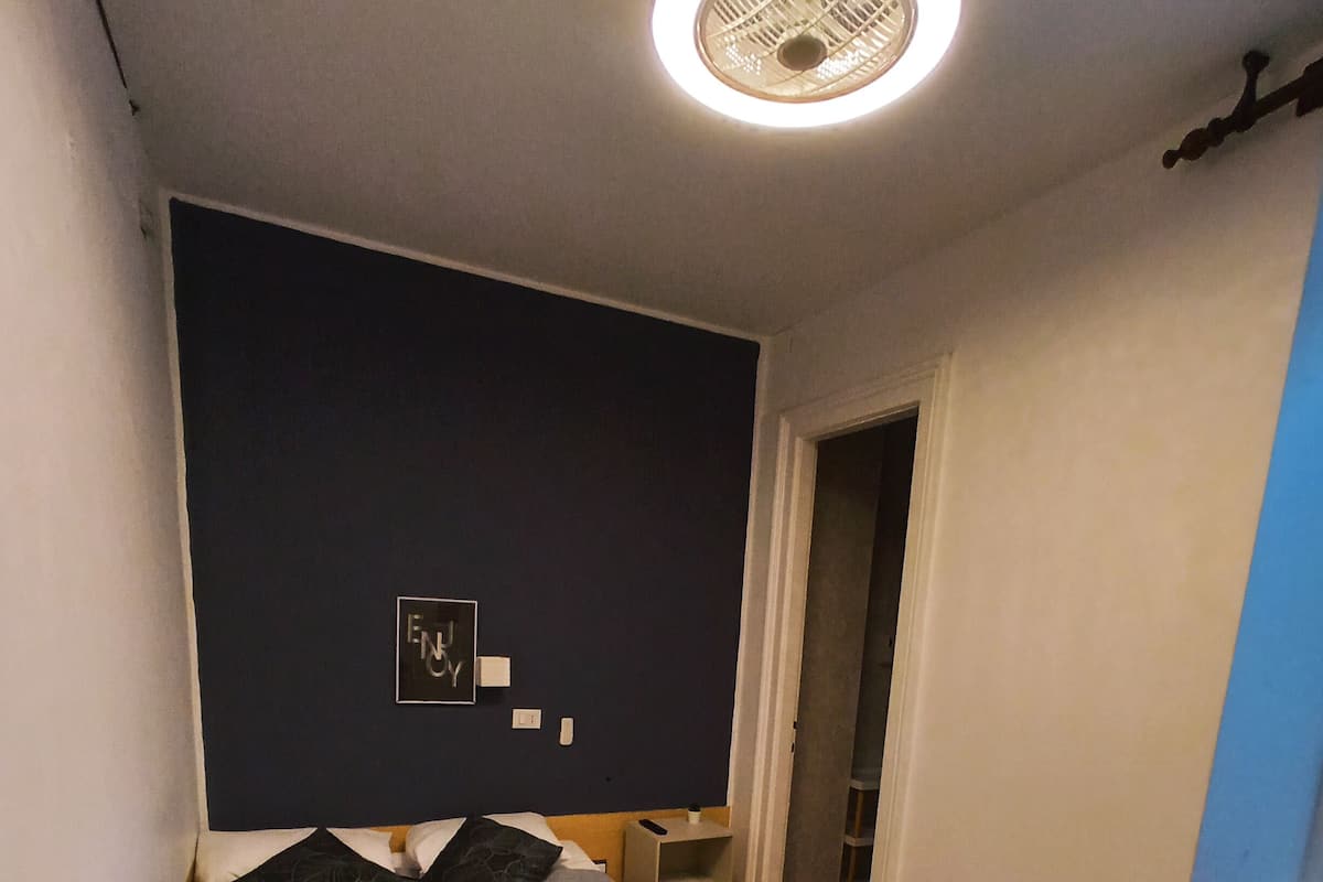 A small, modern bedroom with a bed, a bedside table, a wall-mounted light fixture, and a door leading to another room.